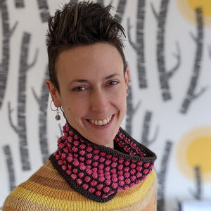 Person facing the camera smiling wearing a pink and grey hand knitted cowl and a yellow striped knitted top
